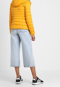 Żółta pikowana kurtka z kapturem, luźne jasnoniebieskie dżinsy w krótkim kroju oraz czarne trampki z białymi akcentami. Widoczne ściegi i struktura materiału.