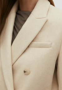 Beige uldblazer med notch lapel, dobbeltradet design og en enkelt brystlomme, der har en rund knapdetalje.