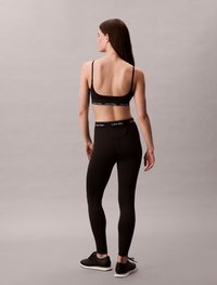 Zwart sportbeha met dunne bandjes, voorzien van een logoband, gecombineerd met hoge zwarte leggings en zwarte sneakers. Gladde textuur.
