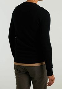 Černý pletený svetr s žebrovanými manžetami a lemem, se strukturovaným vzorem. Nosený přes světle béžovou košili a v kombinaci s kostkovanými kalhotami.