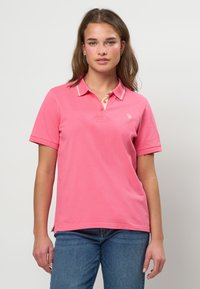 Mlada ženska z rjavimi lasmi, oblečena v roza polo majico s kratkimi rokavi in modre kavbojke, stoji pred enobarvnim svetlim ozadjem.