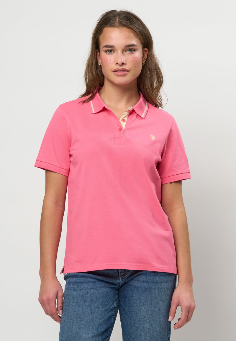 Mlada ženska z rjavimi lasmi, oblečena v roza polo majico s kratkimi rokavi in modre kavbojke, stoji pred enobarvnim svetlim ozadjem.