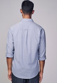 Camicia button-up azzurro chiaro con maniche arrotolate, dettaglio a piega sul retro, orlo arrotondato e texture del tessuto visibile. Adatta per un look casual.