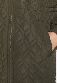 Giacca imbottita verde oliva con motivo a diamante, dotata di cerniera frontale e tasche, realizzata in un materiale morbido e leggero.