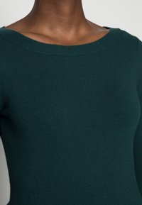 Gros plan sur une personne portant un pull en laine vert foncé avec un col large et arrondi, sur un fond uni.
