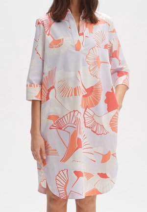 Robe ample blanche avec motifs floraux abstraits corail et orange clair, manches trois-quarts, col en V, portée par une femme.