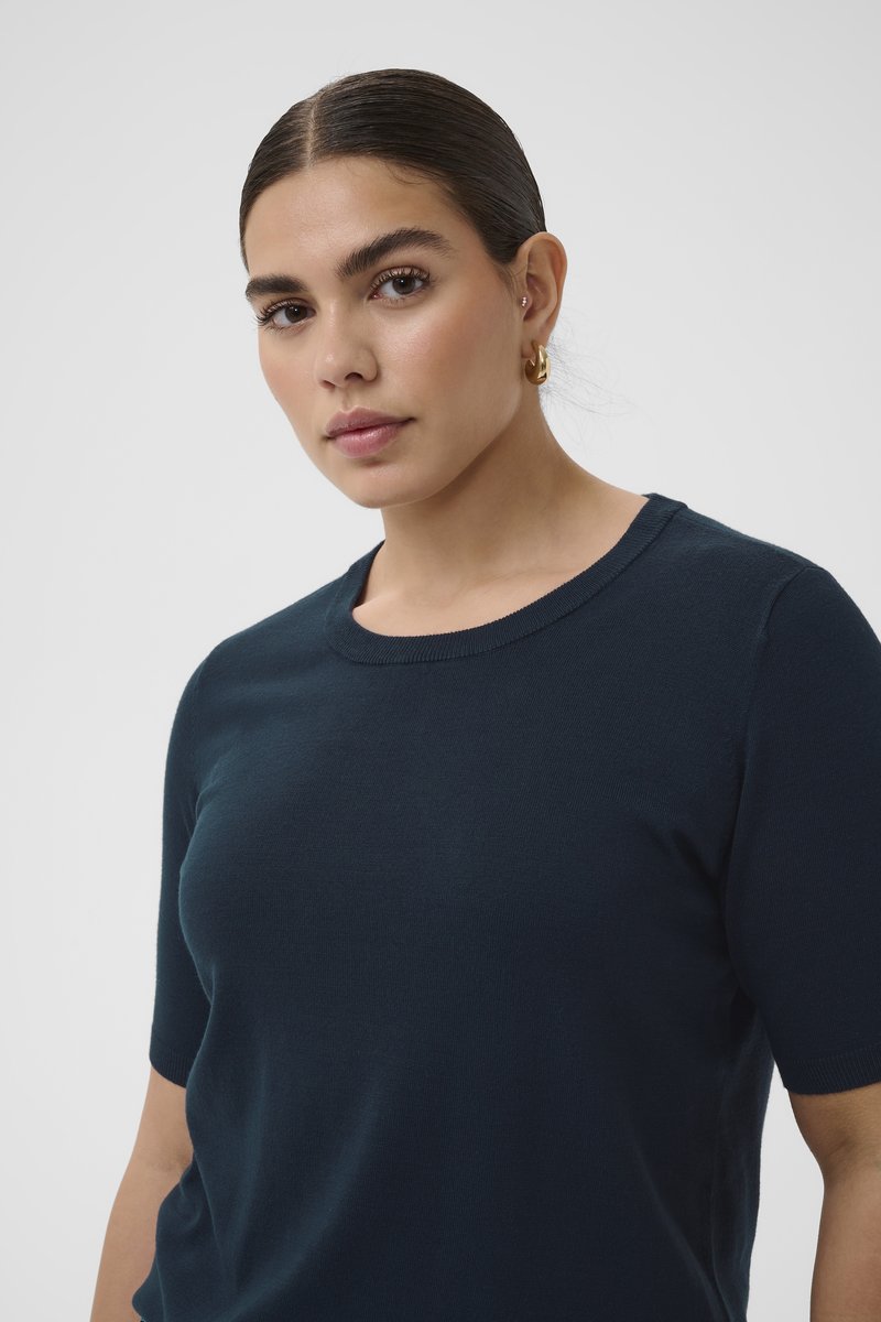 Femme aux cheveux foncés tirés en arrière, portant de petites boucles d'oreilles en or en forme d'anneau et un haut bleu foncé à manches courtes, regardant vers l'avant avec une expression neutre.