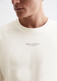 Tej színű póló, puha pamutból készült, kerek nyakkivágással és a "Marc O'Polo" felirattal a mellkas felett.