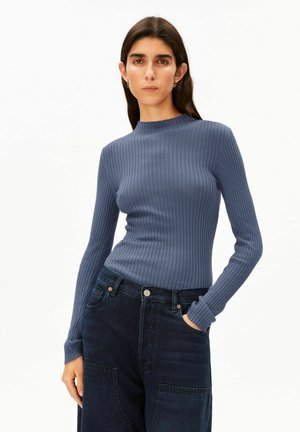 Femme aux cheveux foncés portant un haut bleu côtelé à manches longues ajusté et un jean foncé taille haute, une main dans la poche, sur fond blanc.