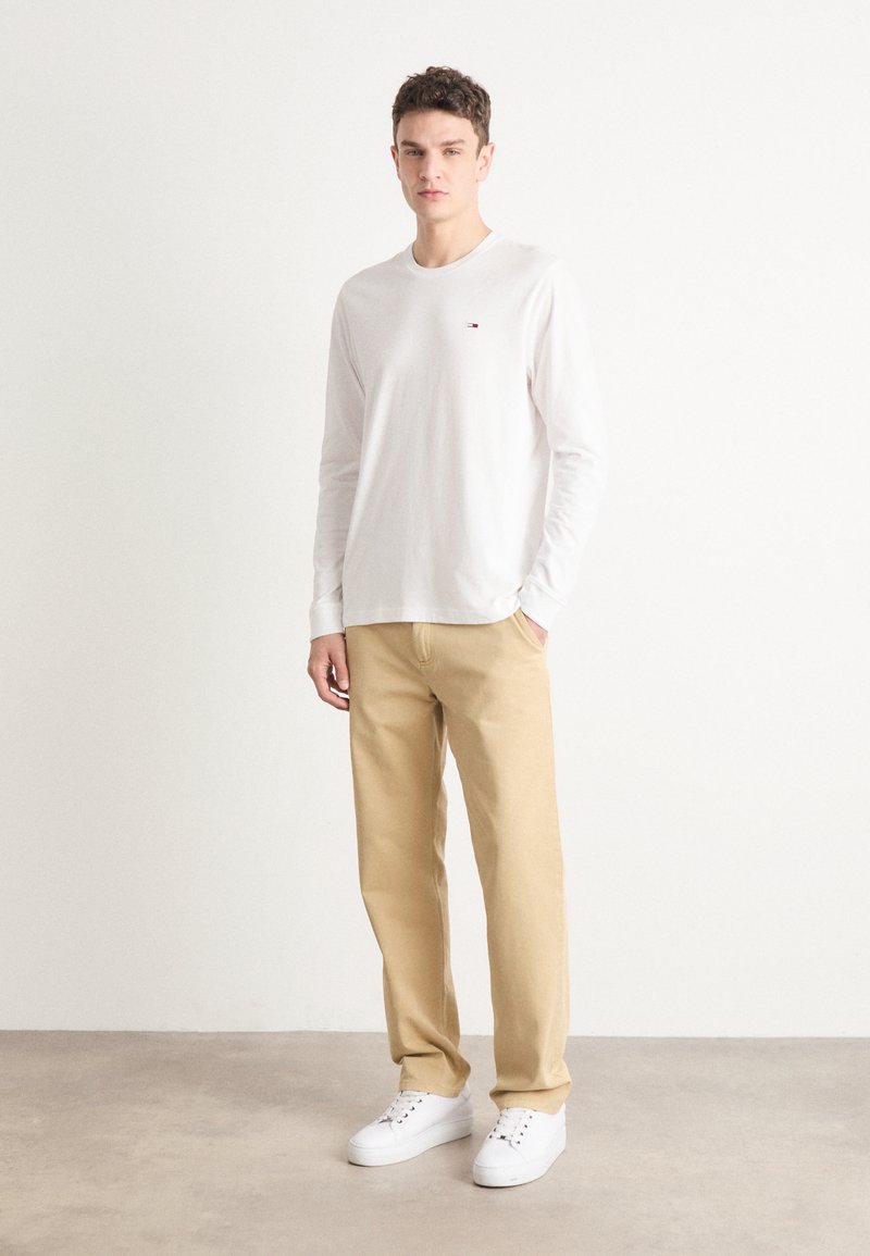 Camiseta blanca de manga larga con un pequeño logo rojo, combinada con pantalones beige y zapatillas blancas, de pie contra un fondo sencillo.