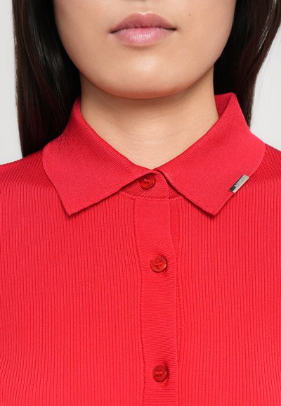 Rød ribbede polo skjorte med knaplukking foran, med matchende knapper og en liten metallisk aksent på kragen.