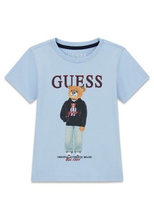 Jasnoniebieska koszulka z krótkim rękawem z niedźwiedziem ubranym w czarną bluzę z kapturem i dżinsy, tekst "GUESS" nad obrazkiem oraz "Original Authentic Brand Est. 1981" poniżej.