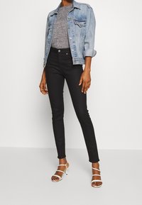 Giacca in denim azzurro chiaro, T-shirt grigia, jeans skinny neri e tacchi bianchi open-toe. Il tessuto è liscio con dettagli minimi.