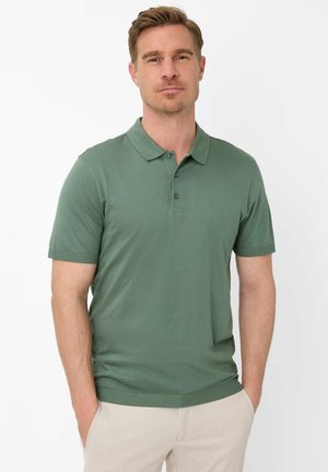 Blonde man die een groen poloshirt met korte mouwen en lichtbeige broek draagt, staat met zijn handen in zijn zakken tegen een witte achtergrond.
