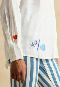 Valge linane särk, mille varrukatel ja äärel on punase ja sinise südamepitsiga tikand, ees nööbiga kinnitused. Sobitatud siniste ja valgete triibuliste pükstega.