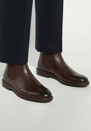 Brown leather Chelsea boots with elastic side panels, a rounded toe, and a low heel, paired with dark trousers. Smooth texture and minimal stitching.