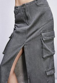 Gonna in denim grigio con spacchi laterali, tasche cargo e vestibilità rilassata. Presenta un chiusura frontale con bottone e cuciture a contrasto.