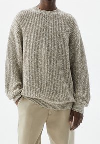Personne portant un pull en maille épaisse beige et blanc, associé à un pantalon beige clair, avec une main dans la poche et un fin collier en chaîne d'argent.