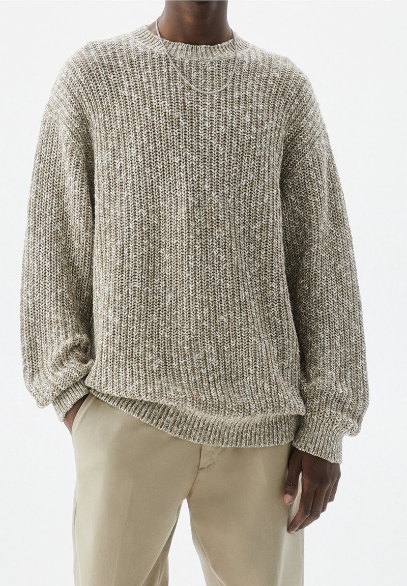 Personne portant un pull en maille épaisse beige et blanc, associé à un pantalon beige clair, avec une main dans la poche et un fin collier en chaîne d'argent.