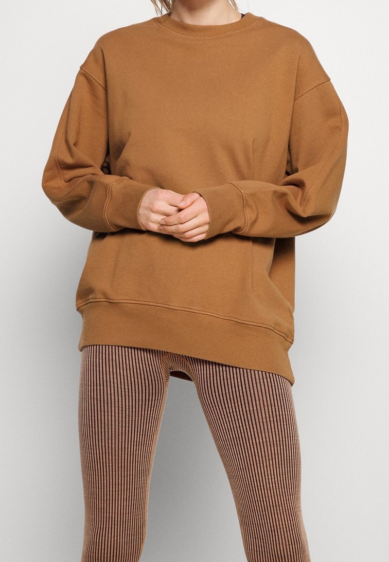 Personne portant un sweat-shirt marron ample avec des poignets côtelés et des leggings rayés verticalement marron et noir, debout devant un fond uni.