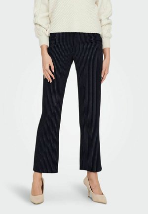 Mujer con pantalones negros de rayas finas de pierna recta combinados con tacones altos beige y un suéter blanco de punto.