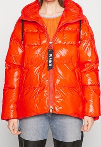 Błyszcząca, puszysta pomarańczowa kurtka z kapturem, z zamkiem błyskawicznym i regulowanymi sznurkami, z widoczną metką marki z przodu.