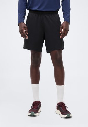 Mann trägt schwarze Sportshorts, weiße Sportsocken, burgunderrote Laufschuhe und ein dunkelblaues Langarmshirt, steht vor einem schlichten Hintergrund.