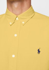 Nära bild av en person som bär en gul skjorta med knappar och en blå broderad polo-spelare logotyp på bröstet.