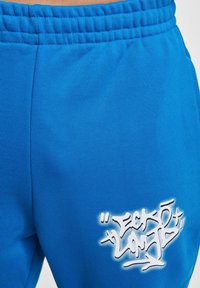 Blauwe sweatpants met een gestructureerde afwerking en elastische tailleband, met een wit graffiti-achtig logo op de linker bovenbeen.