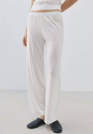 Frau trägt weite weiße Hose, ein weißes bauchfreies Oberteil und schwarze Slip-On-Schuhe, steht auf einem hellen Boden vor einem schlichten Hintergrund.