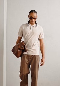 Beige polo skjorte med korte ermer, brune bukser og en brun lærveske. Tilbehøret inkluderer mørke solbriller. Enkel, elegant design.