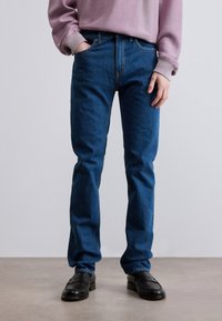 Μπλε τζιν παντελόνι με ίσια γραμμή, διαθέτει κλασικό σχεδιασμό με πέντε τσέπες και έντονη ραφή σε αντίθεση. Φορώντας με μαύρα παπούτσια.