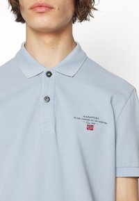 Polo bleu clair avec un col classique et deux boutons noirs. Présente un logo et un texte brodés sur la poitrine. Tissu en mélange de coton.