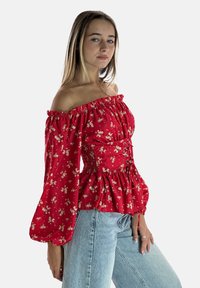 Blusa rossa off-the-shoulder con stampa floreale, corpino arricciato e maniche lunghe a sbuffo. Abbinata a jeans azzurri.