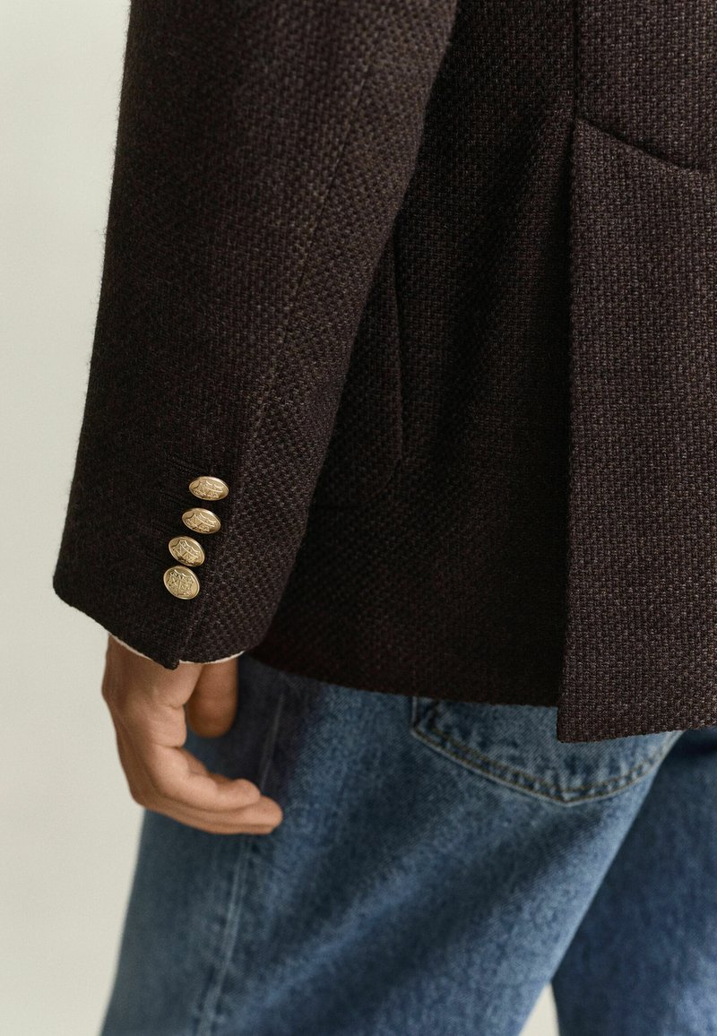 Person wearing a dark textured blazer with four gold buttons on the sleeve and blue jeans, shown from mid-torso down.