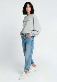 Felpa grigia con testo nero, vestibilità morbida, abbinata a jeans a vita alta di colore blu chiaro. Il modello indossa sneaker bianche, in piedi su uno sfondo neutro.