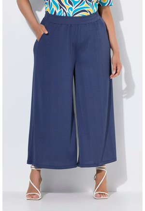 Femme portant un pantalon large bleu marine avec poches latérales et des sandales blanches à talons hauts à brides, debout devant un fond clair.