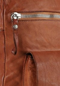 Dettaglio di borsa in pelle marrone con cerniera metallica argentata, superficie testurizzata e anello in pelle. Design minimalista con cuciture visibili.