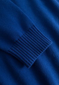 Détail d'un pull bleu tricoté avec un poignet côtelé, une texture douce et un tissu lisse, mettant en valeur les coutures régulières et la couleur vive.