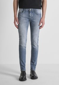 Muškarac u izbijeljenim svijetloplavim uskim trapericama i crnim kožnim čizmama stoji nasuprot običnoj bijeloj pozadini.