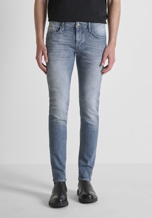 Hombre con jeans ajustados de un azul claro desgastado y botas de cuero negras, de pie contra un fondo blanco liso.