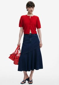 Cardigan rosso a maniche corte con bottoni, abbinato a una gonna a linea A in denim scuro. Porta una borsa rossa. Sandali blu con cinturini e dettagli floreali.