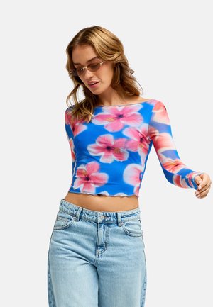 Kobieta nosząca brązowe okulary przeciwsłoneczne, niebieski crop top z długim rękawem z różowymi motywami kwiatowymi i jasnoniebieskie dżinsy z wysokim stanem.