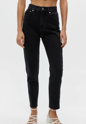 Woman wearing high-waisted black jeans and white strappy block-heel sandals, shown from waist to feet against a plain background.