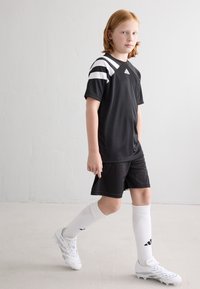 Tenue de sport noire comprenant un t-shirt à manches courtes avec des rayures blanches, associé à un short et des chaussettes blanches, porté avec des chaussures de sport blanches.