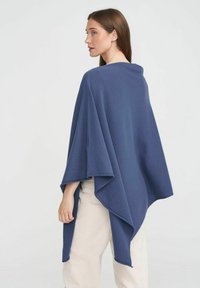 Marineblå, mjuk tygponcho med en lös och draperad design. Har breda ärmar och en rundad nederkant, bärs över ljusa byxor.