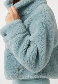 Světle modrá plyšová bunda s vysokým límcem a texturovanou tkaninou. Na lemu má trojúhelníkový kovový dekorativní prvek s logem.