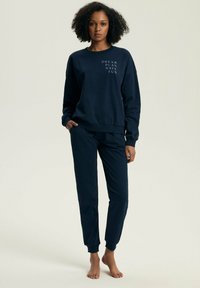 Granatowy sweter z długim rękawem i dopasowane spodnie z miękkiej, teksturowanej tkaniny. Sweter ozdobiony jest białym napisem: "MARZ, PLANUJ, BAW SIĘ". Spodnie mają elastyczne mankiety.