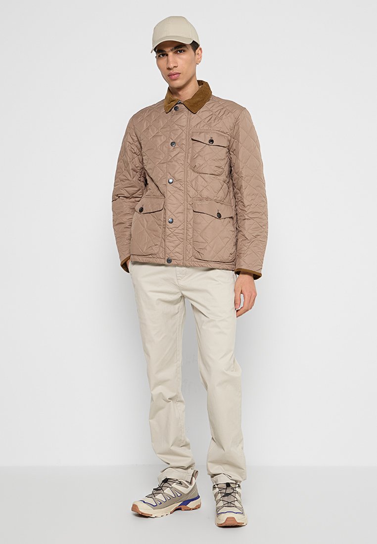 Veste matelassée marron avec un col en velours côtelé marron, deux poches poitrine et fermeture à boutons ; associée à un pantalon beige et des chaussures de couleur claire.