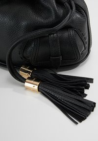 Sac en cuir noir avec un matériau texturé, un design décontracté et du matériel en métal doré, agrémenté de pompons en cuir noir bien visibles.